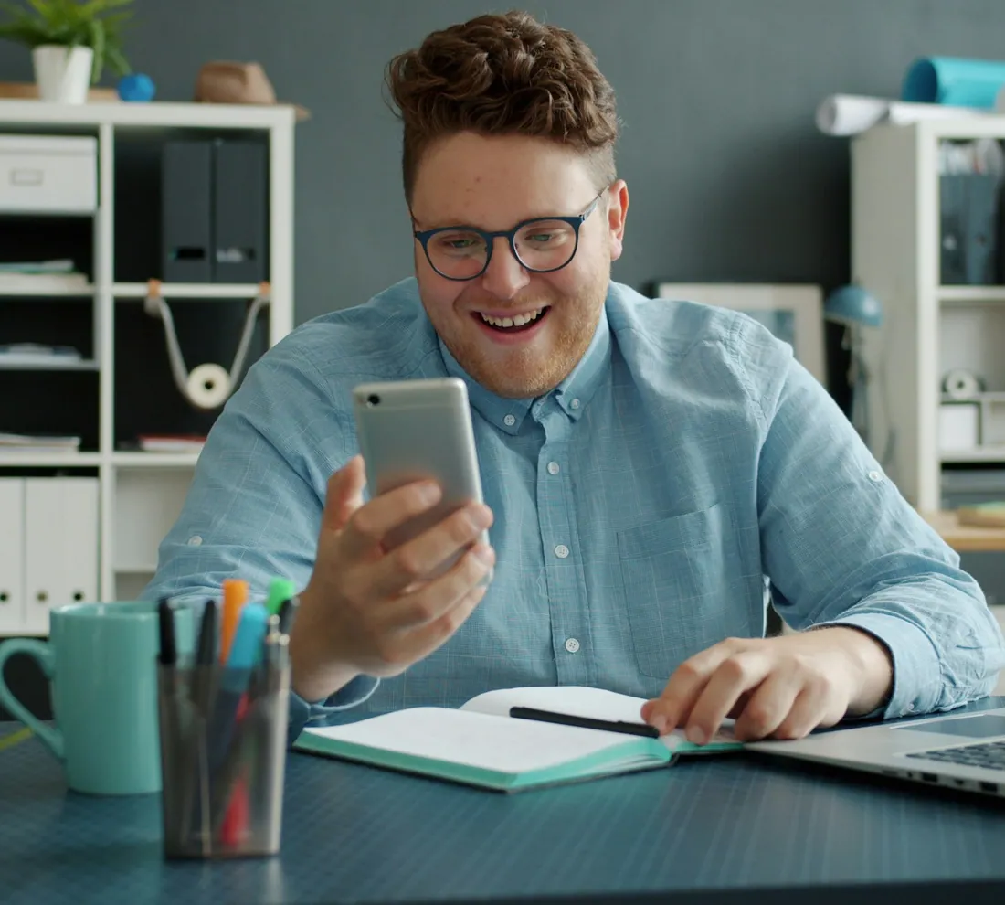 Homem sorrindo enquanto conversa pelo celular, sentado à mesa com o notebook